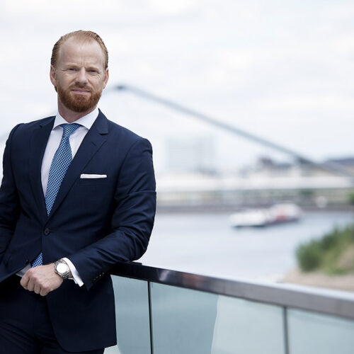 Exklusives Business-Portrait: Mann im blauen Anzug posiert vor einer Stadtkulisse am Wasser.