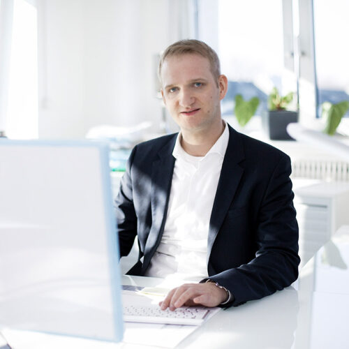 Business-Portrait Essen: Manager im dunklen Sakko vor einem Büroarbeitsplatz.