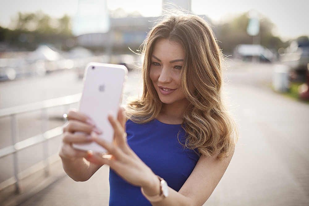 Werbefotografie weibliche Person im blauen Kleid mit langen braunen Haaren macht ein Selfie mit einem Apple Smartphone