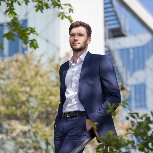 Business-Lifestyle-Portrait: Eleganter Mann im blauen Anzug posiert draußen vor moderner Architektur.